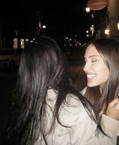 two women standing next to each other on a street at night with lights in the background