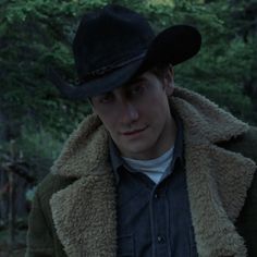 a young man wearing a black hat and coat in the woods with trees behind him