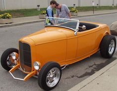 two men standing next to an orange car on the street