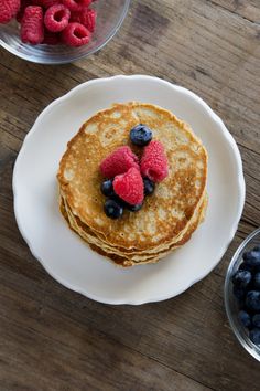 pancakes with berries and blueberries sit on a white plate next to bowls of raspberries