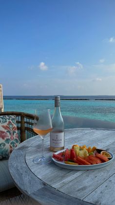 a bottle of wine and plate of fruit on a table with water in the background