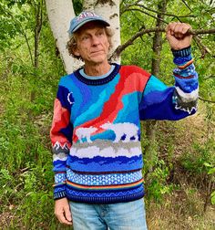 an older man standing in front of a tree wearing a colorful sweater and hat with his fist raised