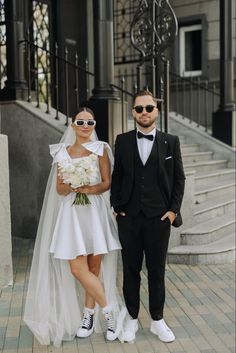 a bride and groom standing on the sidewalk in front of an old building wearing sunglasses