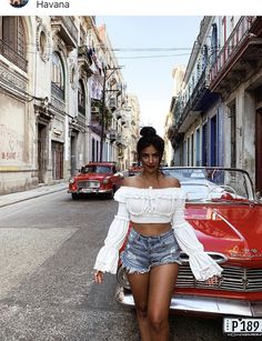 a woman walking down the street in front of a red car with her hands on her hips