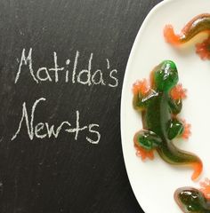 a green and orange frog sitting on top of a white plate next to a chalkboard