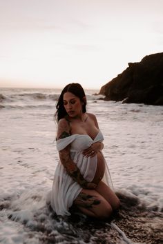 a pregnant woman sitting in the water at the beach with her hands on her stomach