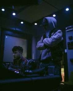 two men standing in a dark room looking at something on the computer screen and one is wearing a hoodie