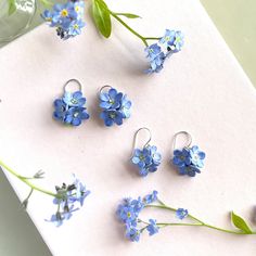 blue flowers are sitting next to some silver earring sets on a white surface with green leaves