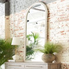 a white dresser sitting next to a large mirror on top of a wooden table with a plant in front of it