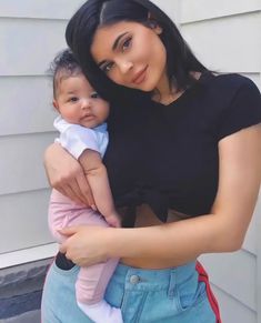 a woman holding a baby in her arms and looking at the camera while standing next to a house