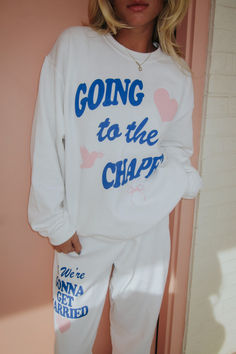 a woman standing in front of a pink wall wearing a white sweatshirt and sweatpants