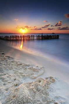 the sun is setting over an ocean with sand on the shore and water in the foreground