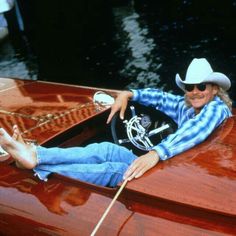 a man wearing a cowboy hat and sunglasses sitting in a red boat with his hand on the steering wheel