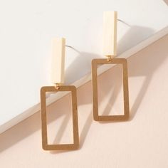 two square shaped gold earrings on top of a white surface with a light pink wall in the background