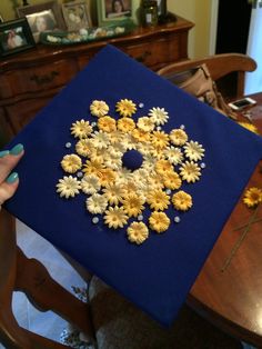 a person holding up a blue and yellow square with daisies on the inside, in front of a dining room table