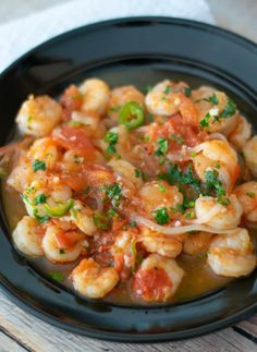 a black plate filled with shrimp and vegetables on top of a wooden table next to a napkin