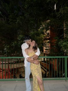 a man and woman embracing each other in front of a green fence with trees behind them