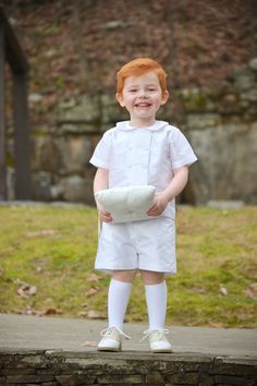 a little boy dressed in white is standing on a stone wall and smiling at the camera