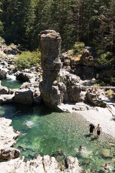 two people are wading in the water near some large rocks and trees, while another person is standing next to them