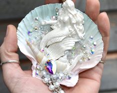 a hand holding a white shell with a mermaid on it's side and pearls in the middle