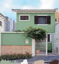 a green house with a white gate and trees in front of it, surrounded by tall buildings