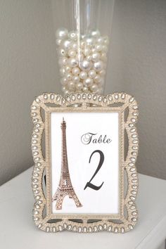 a table number frame with pearls in front of the eiffel tower and a vase filled with flowers