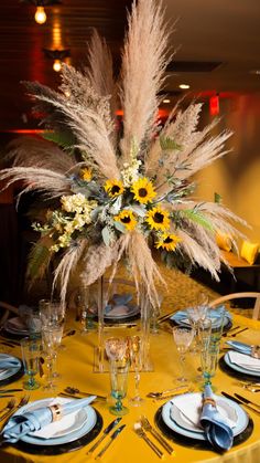 a table set with blue plates and silverware, yellow napkins and tall centerpiece