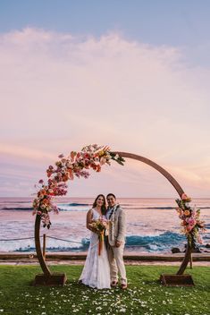 a bride and groom standing under an arch with flowers on the grass near the ocean