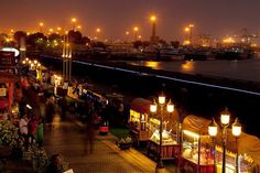 an outdoor market with lots of lights and people walking around it at night near the water