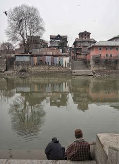 two people sitting on the edge of a body of water with buildings in the background