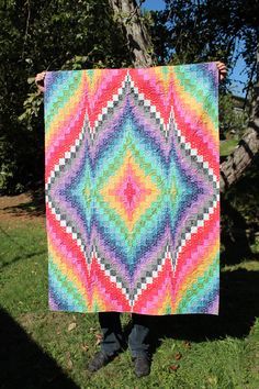 a woman holding up a colorful quilt in front of a tree with grass and trees behind her