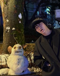 a man sitting on the ground next to a snowman in front of a tree