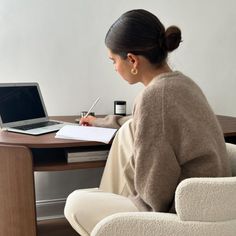 a woman sitting at a desk with a laptop computer and pen in her hand, writing