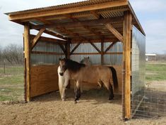 two horses are standing under a wooden shelter in a field with grass and dirt on the ground