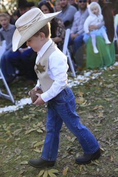 a young boy wearing a cowboy hat and jeans walking through the grass with people in the background