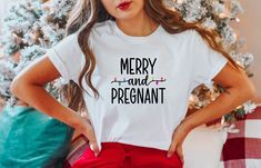 a woman standing in front of a christmas tree wearing a merry and pregnant t - shirt