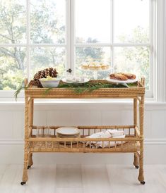 a wicker serving cart with food on it in front of a window and pine cones