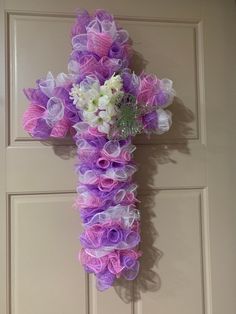 a cross made out of purple and white flowers hanging on the front door with ribbon