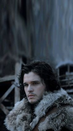 a man with long hair and a fur collar standing in front of a wooden structure