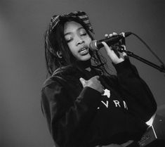 a woman singing into a microphone while holding a guitar in front of her face and wearing a hoodie