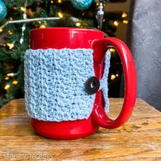 a crocheted coffee cup cozys up in front of a christmas tree with a red mug holder