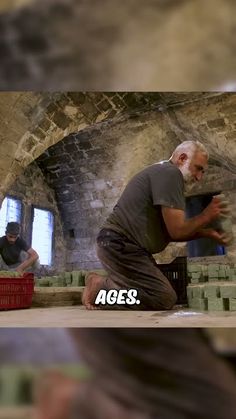 an old man kneeling down in front of a brick wall with another man standing next to him