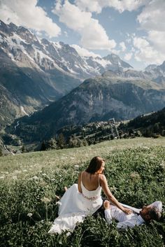 a bride and groom sitting on the grass in front of mountains with snow capped peaks