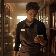 a man in an apron looking at his cell phone while standing in front of bookshelves