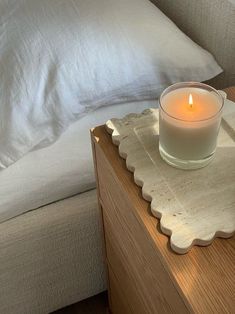 a lit candle sitting on top of a wooden table next to a white bed and pillows