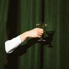 a person holding a wine glass in front of a green curtain with a shadow on it