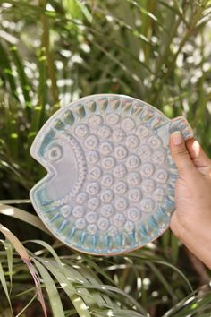 a hand holding a ceramic fish dish in front of some plants