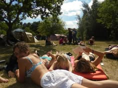 three women laying on the ground in front of tents and trees, one reading a book