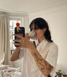 a woman drinking from a cup while standing in front of a bed and holding a cell phone