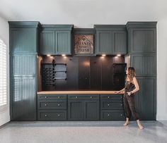 a woman standing in the middle of a kitchen with dark green cabinets and black appliances